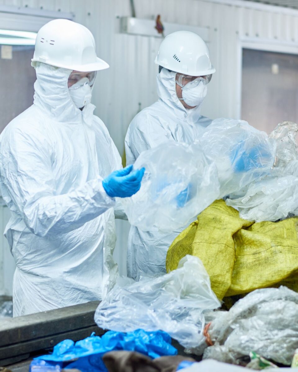 Portrait of two workers wearing biohazard suits at waste processing plant sorting recyclable plastic and cardboard on conveyor belt